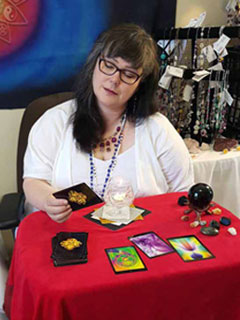 Eileen, Soul To Soul Medium, at a red table cloth covered talble with oracle cards and a glass of water for scrying performing a mediumship reading.