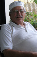 Image of Angel, Cuban Shaman. He is wearing a white short sleeve shirt with a collar and a white hat. He has short wiry white hair, a mustash and is wearing glasses