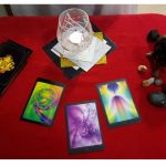 Image of tools a Psychic or Clairvoyant Medium may use. It includes oracle cards, a glass with a candle lit inside of it, a black crystal ball, and some crystals all placed on a table with a red cloth.