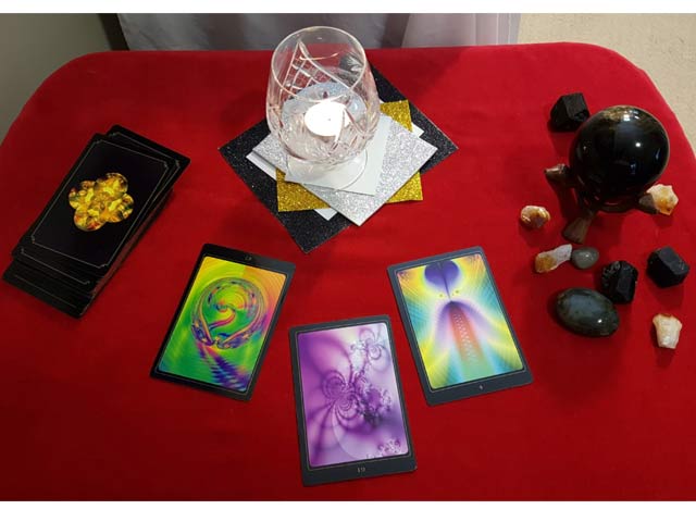 Image of tools a Psychic or Clairvoyant Medium may use. It includes oracle cards, a glass with a candle lit inside of it, a black crystal ball, and some crystals all placed on a table with a red cloth.